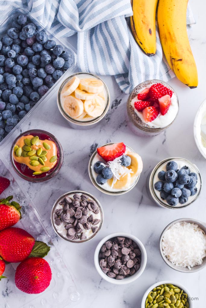 Looking down at mason jars of chia pudding flavors with small bowls of chocolate chips, shredded coconut, seeds, fresh strawberries, blueberries and bananas beside it.