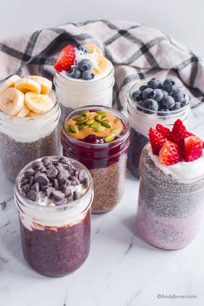 Six chia pudding flavors in mason jars including chocolate, strawberries and cream, pb & j, blueberry, vanilla and banana.