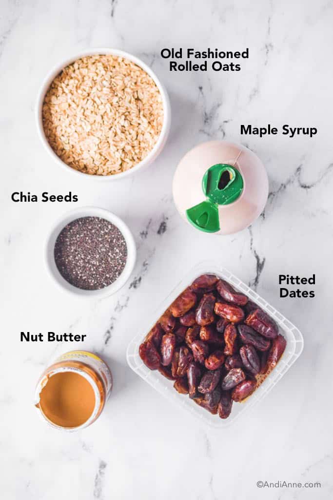 Ingredients on marble counter: bowl of rolled oats, maple syrup, bowl of chia seeds, pitted tates container, jar of nut butter.