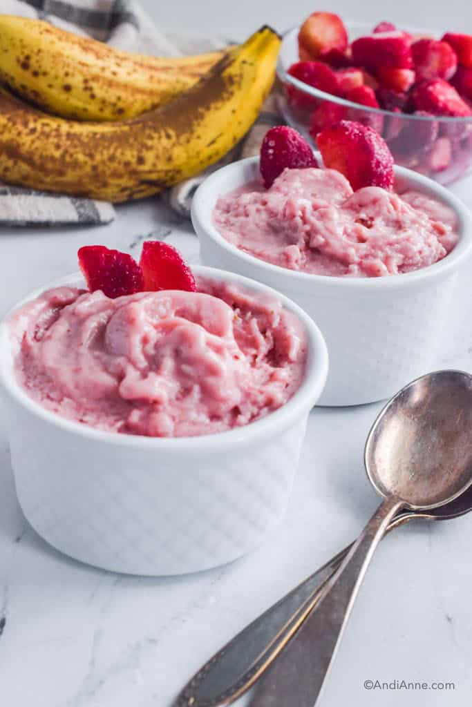 strawberry banana nice cream in white bowls with bananas and frozen strawberries in background. Spoons in foreground.