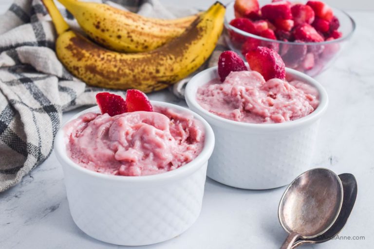 strawberry banana nice cream in white bowls with spoons, ripe bananas, bowl of frozen strawberries and kitchen towel surrounding bowls