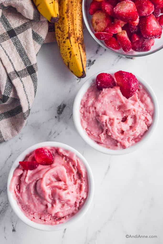 Looking down at strawberry banana nice cream in white bowls with bananas beside them.