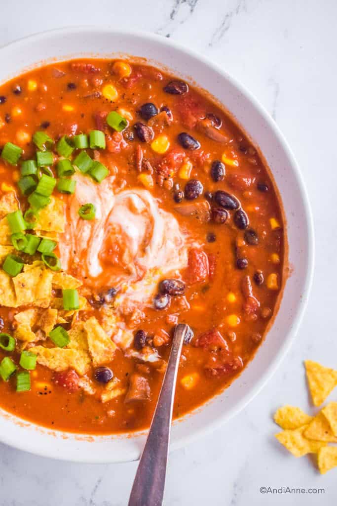 vegetarian tortilla soup in white bowl with spoon. Green onion and crushed tortilla chips sprinkled on top of soup.