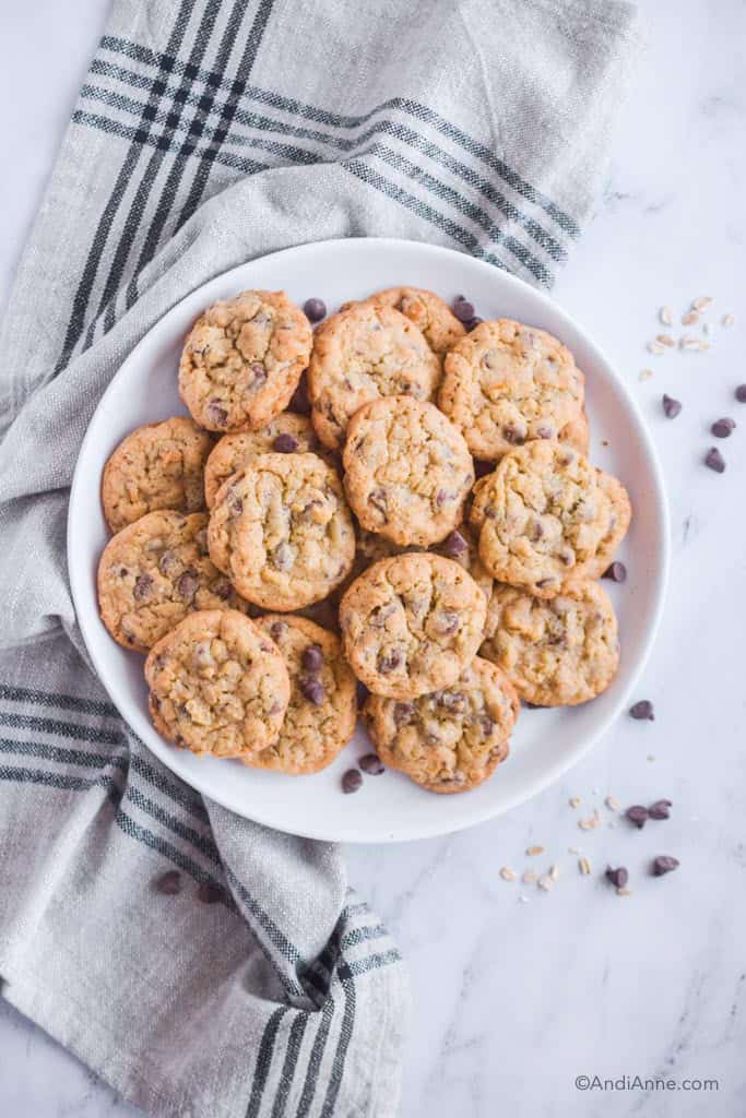 pile of chocolate chip oat cookies on white plate with kitchen towel underneath.