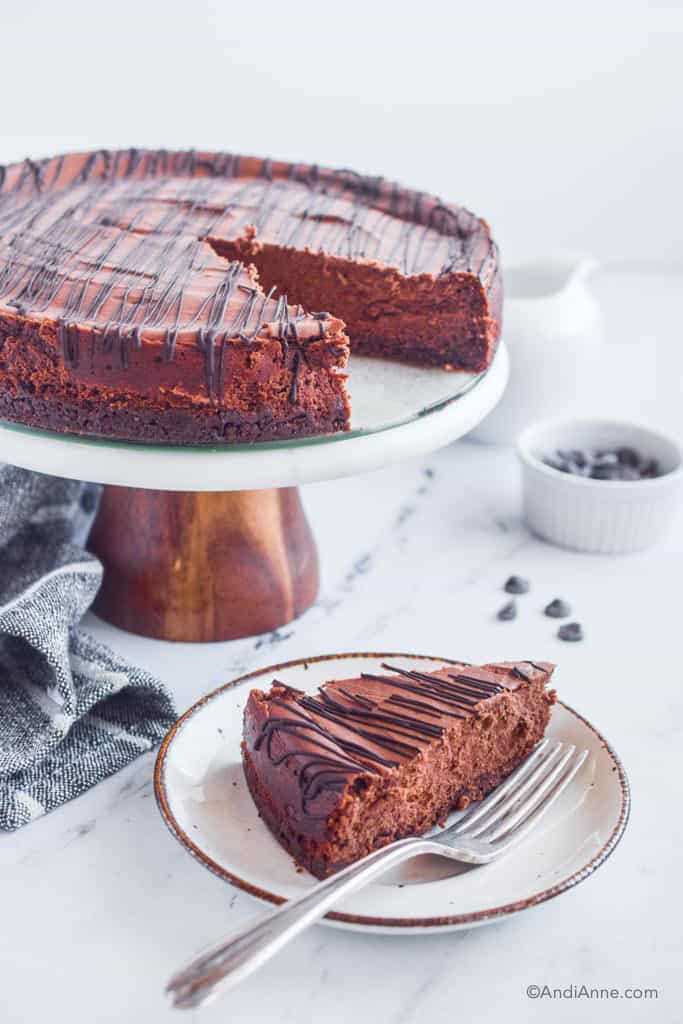Double chocolate cheesecake on cake stand with slice and fork on a plate in front. Small bowl of chocolate chips in background.