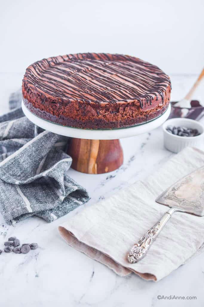 Double chocolate cheesecake on cake stand, kitchen towels, silver pie server, bowl of chocolate chips surround the cake.