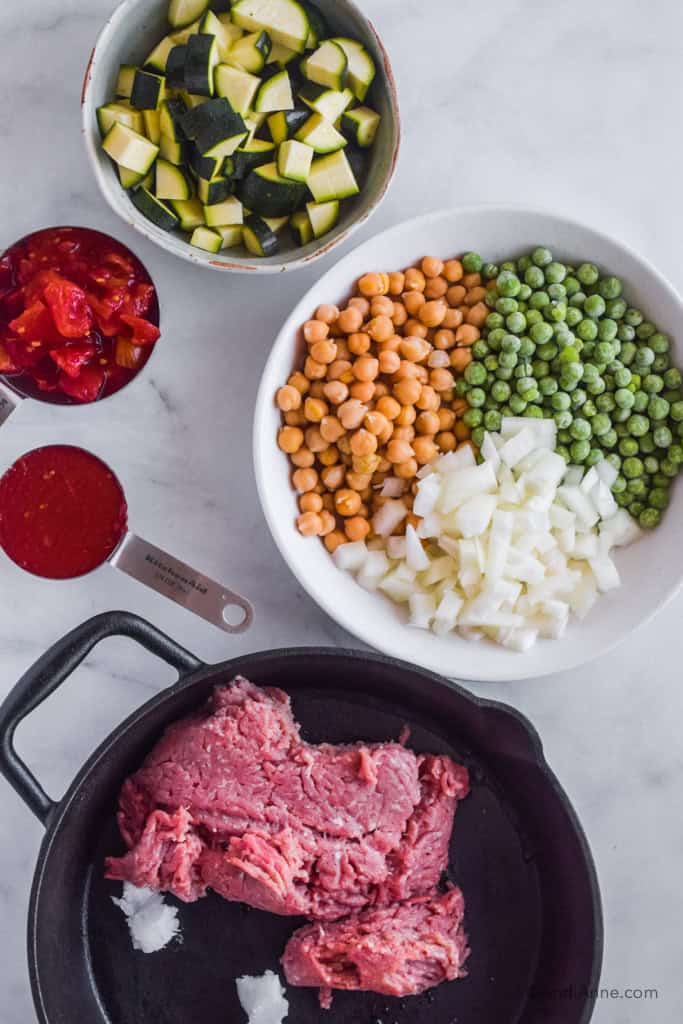 Bowl of chopped zucchini, white bowl of chickpeas, peas, and chopped onion, measuring cup of diced tomatoes, cup of tomato paste, frying pan with raw ground turkey and chunks of coconut oil.