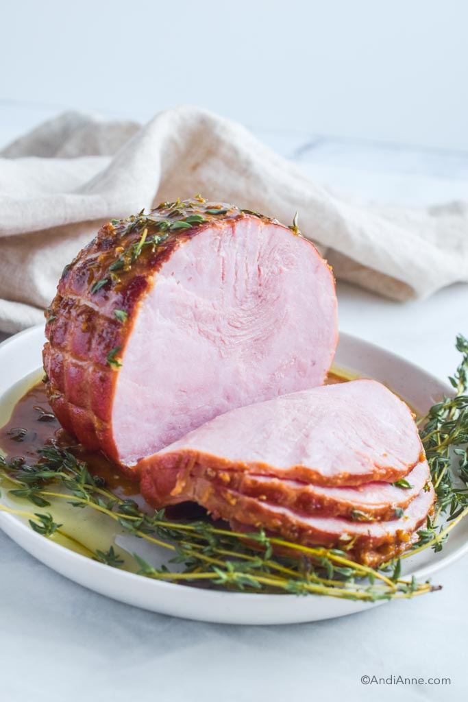 Cooked ham with several slices cut off, sprigs of thyme and honey mustard glaze on a white plate. 