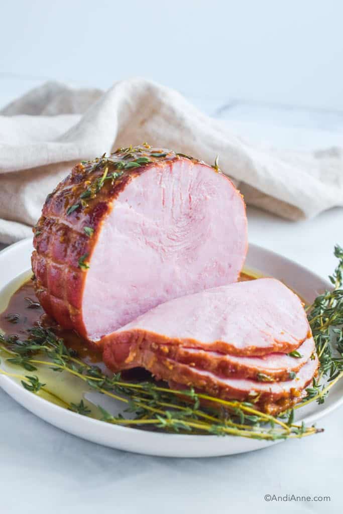 Cooked ham with several slices cut off, sprigs of thyme and honey mustard glaze on a white plate.