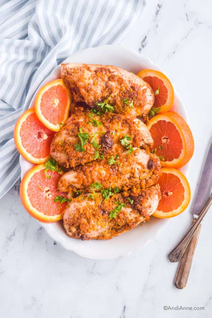 Looking down at a plate with four orange ginger chicken breasts, slices of orange and parmesan sprinkled on top. A knife and kitchen towel are beside the plate.