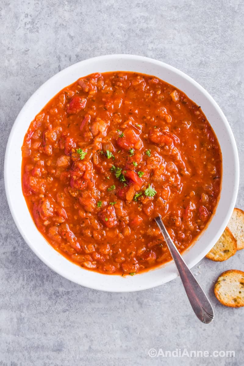 Looking down at white bowl of tomato garlic soup with spoon inside and crackers beside bowl.