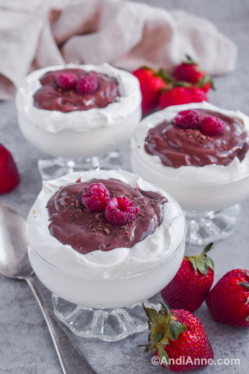 Three cups of pudding in a cloud dessert with fresh raspberries on top and surrounding the cups along with strawberries.