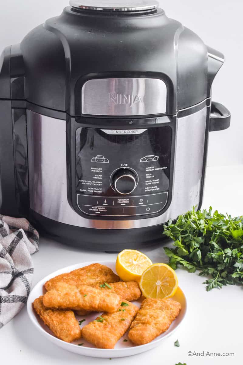 Air fryer frozen fish sticks: Ninja foodi deluxe air fryer with fresh parsley, kitchen towel, and plate of cooked fish sticks and sliced lemons in front.