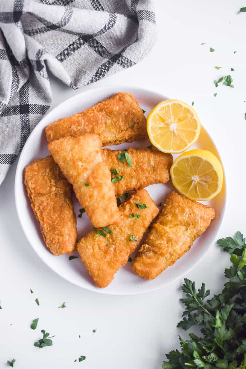 Six fish sticks on a white plate with lemon sliced in half. Kitchen towel and parsley are beside the plate.