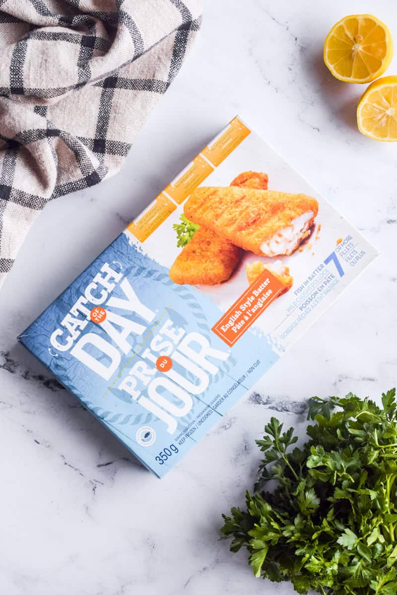 Box of store-bought frozen fish sticks with kitchen towel, fresh parsley and sliced lemon surrounding it.