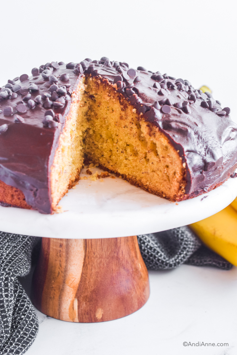 Slice out out of banana cake with chocolate peanut butter frosting and chocolate chips sprinkled on top. Sitting on a cake stand with a kitchen towel behind it. 