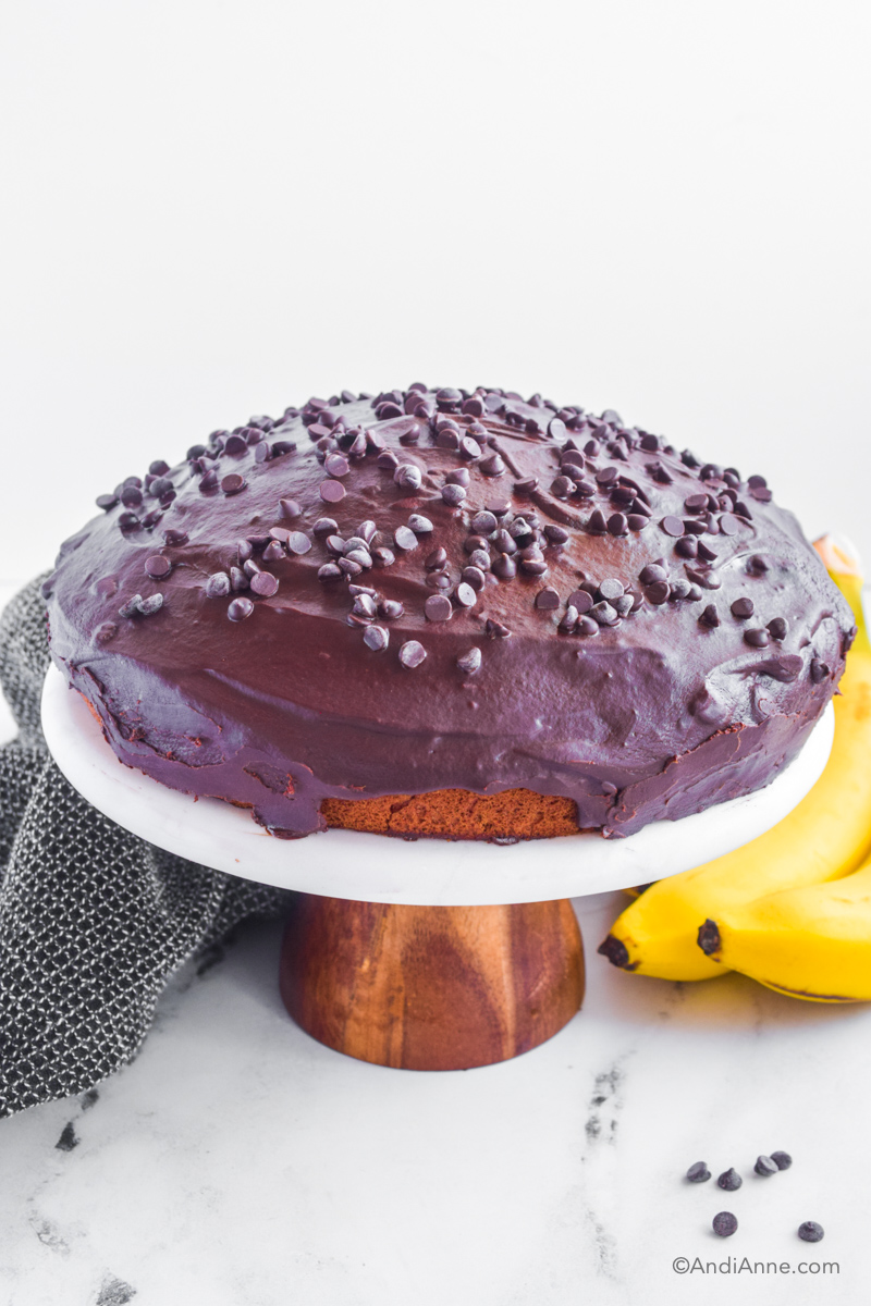 Cake with chocolate frosting and chocolate chips on top. Bananas and kitchen towel behind it. 