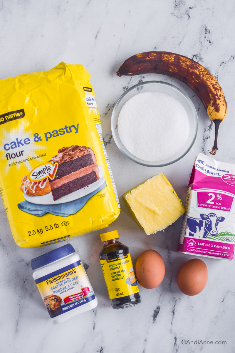 Looking down at ingredients including bag of cake flour, bowl of sugar, over ripe banana, carton of milk, butter, jar of baking powder and vanilla extract, two eggs.