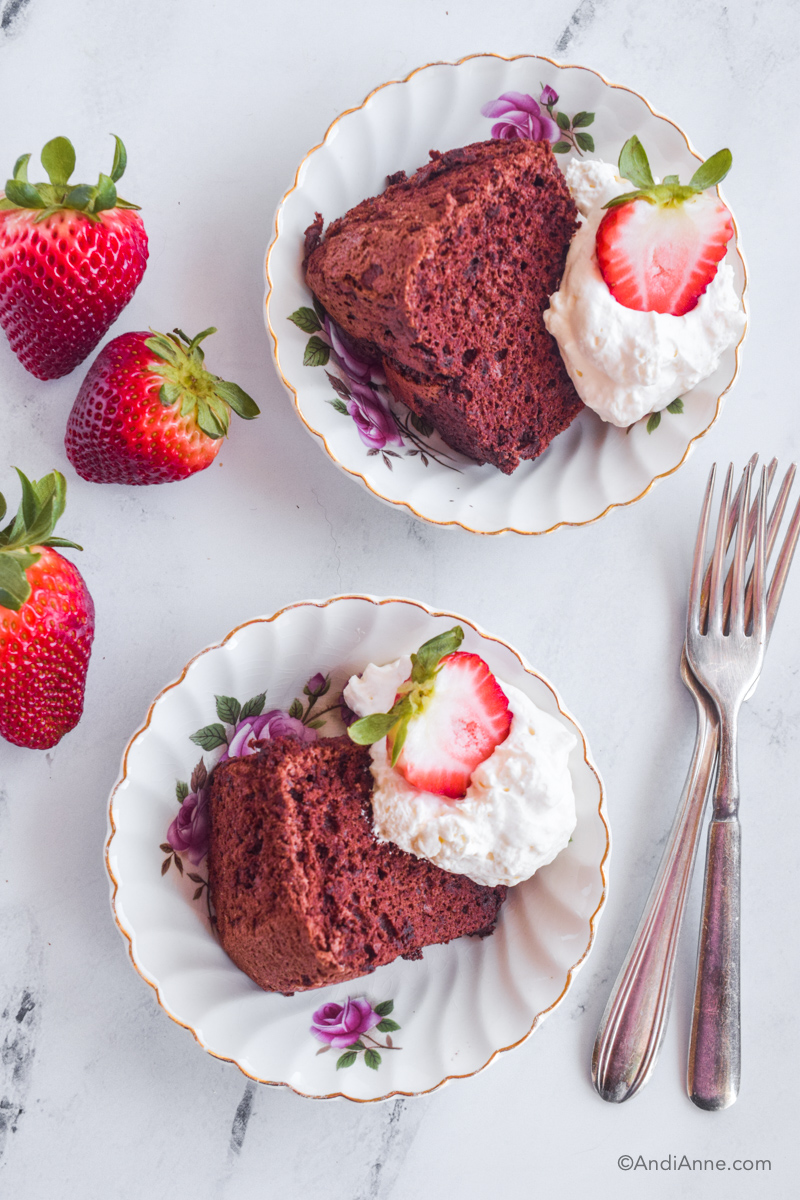 Looking down at two plates with slices of cake, whipped cream and sliced strawberries on a counter. Two forks and three strawberries are beside the plates.