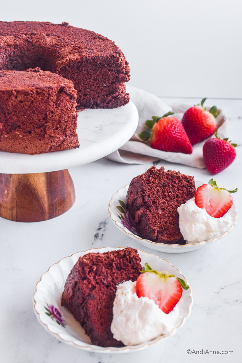 Chocolate cake on cake stand with cake slice, whipping cream and sliced strawberry on a small plate in front. Three strawberries are in the background. 