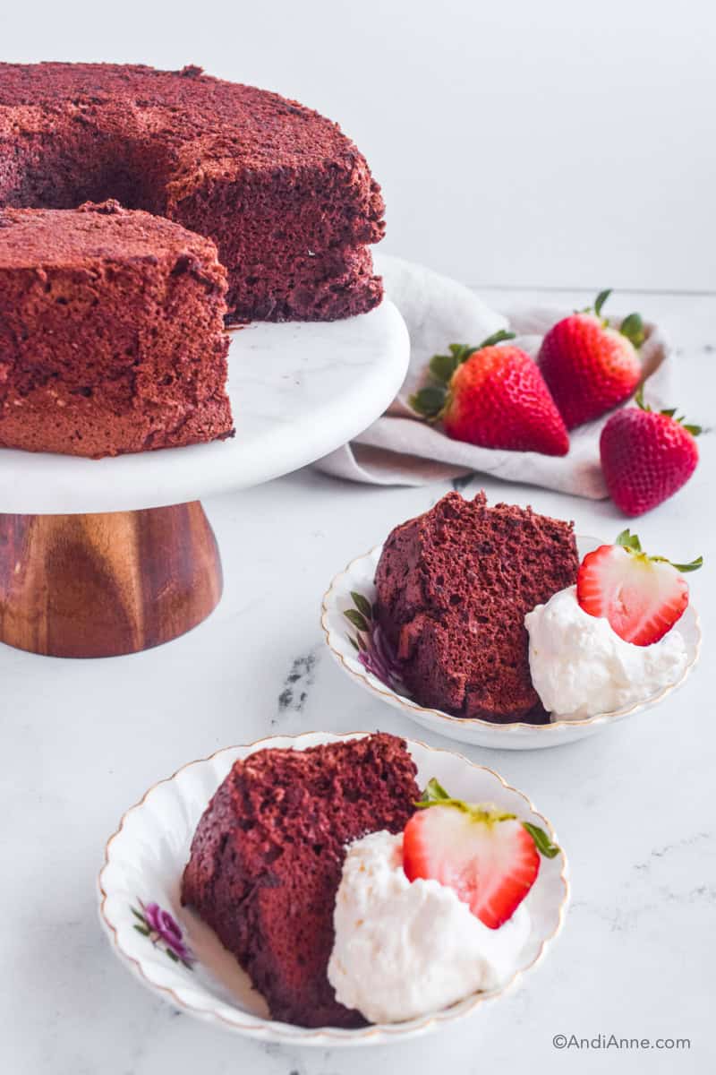 Chocolate cake on cake stand with cake slice, whipping cream and sliced strawberry on a small plate in front. Three strawberries are in the background.