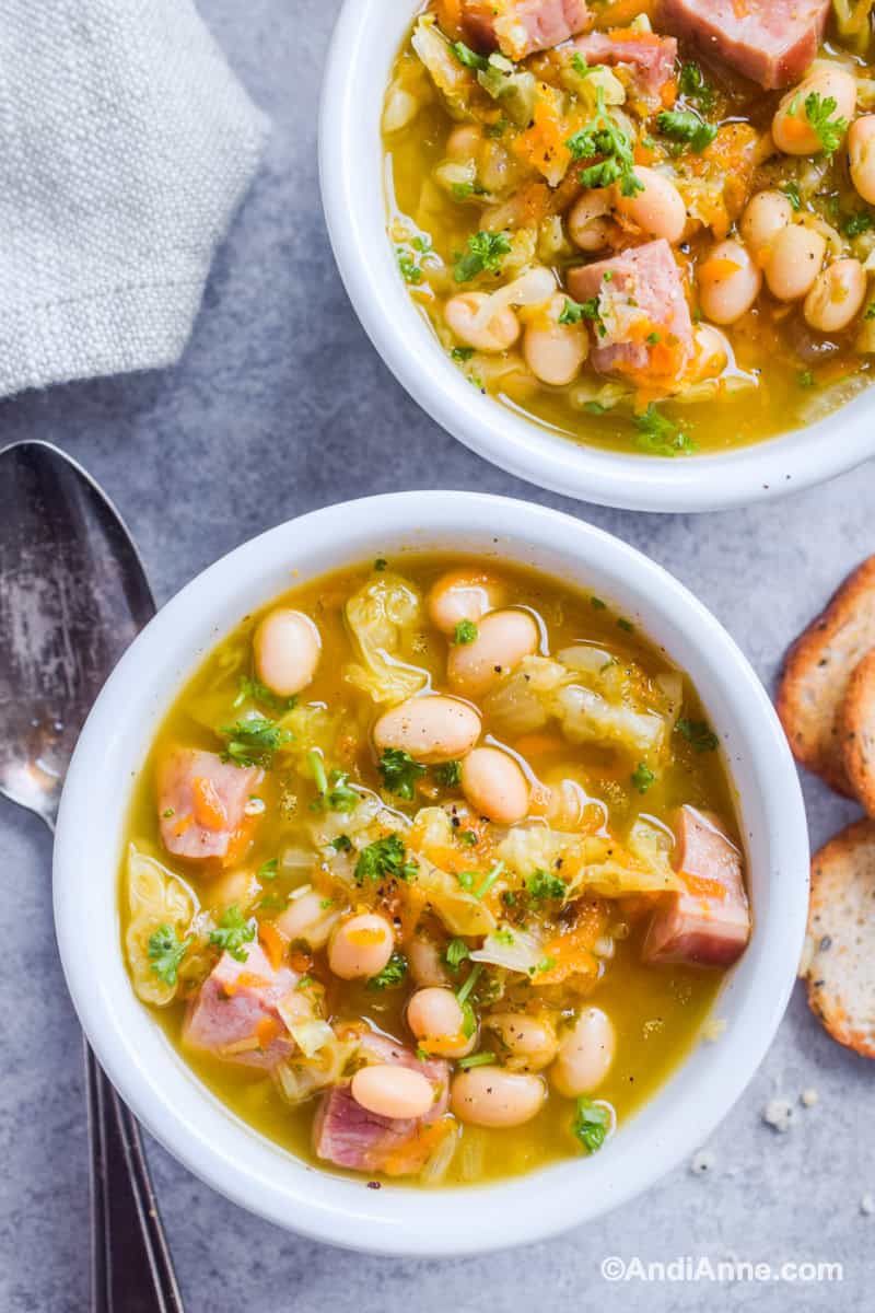 Close up of two white bowls with ham and bean soup with crackers and spoon surrounding it.