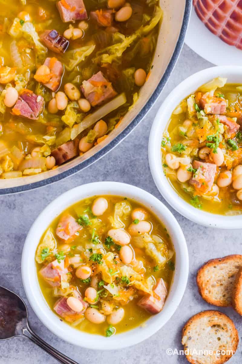 Two bowls of ham and bean soup with pot and crackers beside them.