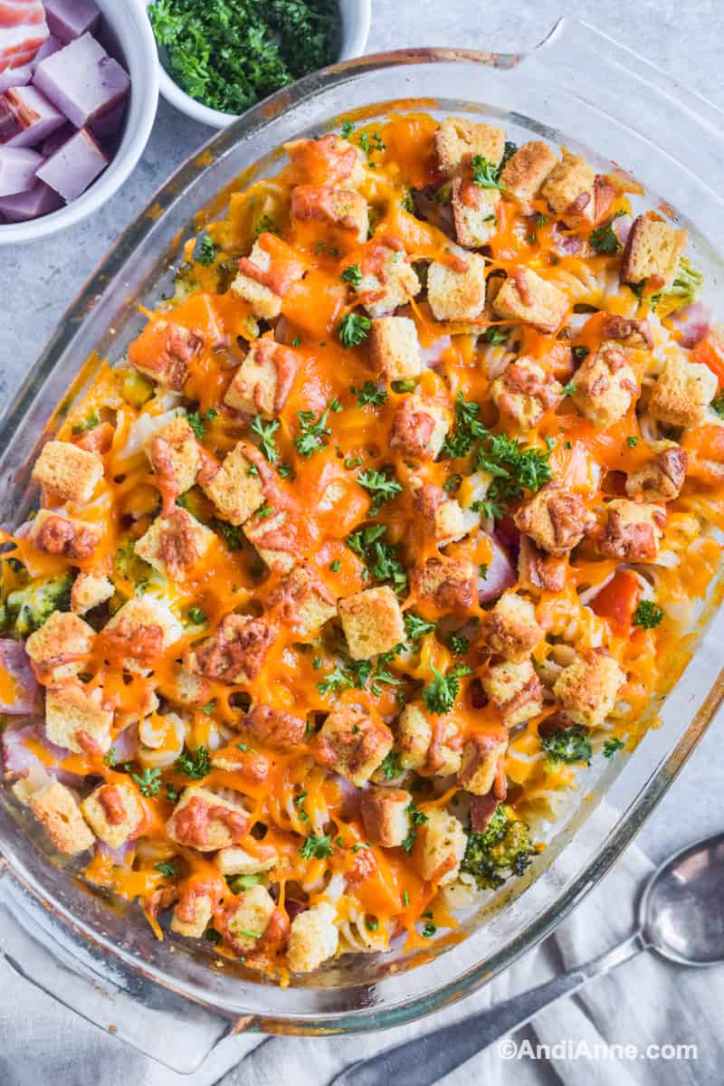 Ham cheese pasta casserole in a glass dish with small bowls of ingredients and a spoon surrounding it.