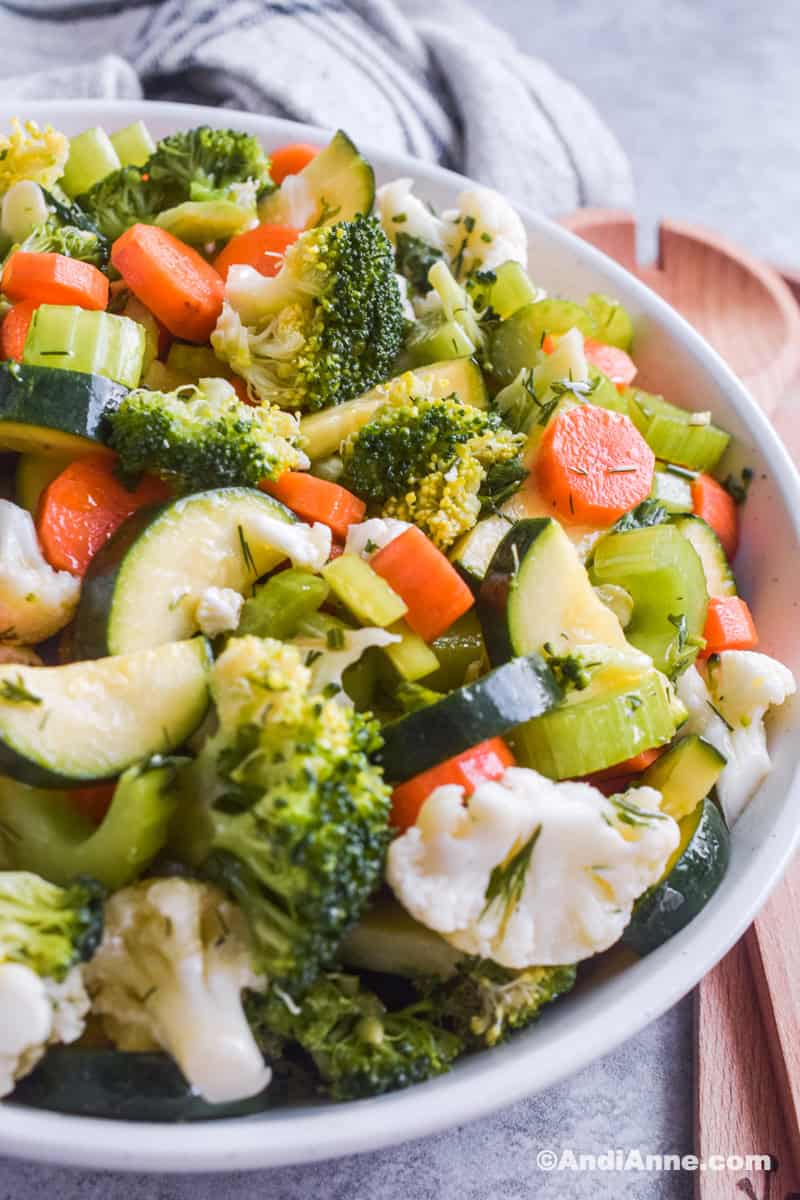Close up of marinated vegetables in a white bowl with salad spoons and kitchen towel in background.