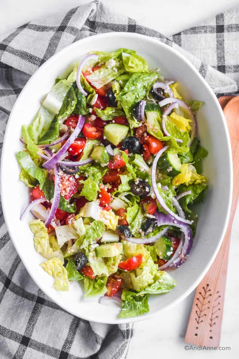 Greek salad with lettuce, onion, cucumber, tomatoes, olives and feta in a large oval bowl with wood salad spoons beside it.