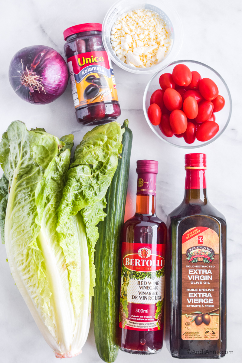 Ingredients lying down on a counter: romaine lettuce, cucumber, red wine vinegar, olive oil, bowl if grape tomatoes, container feta cheese, red onion, container of olives. 
