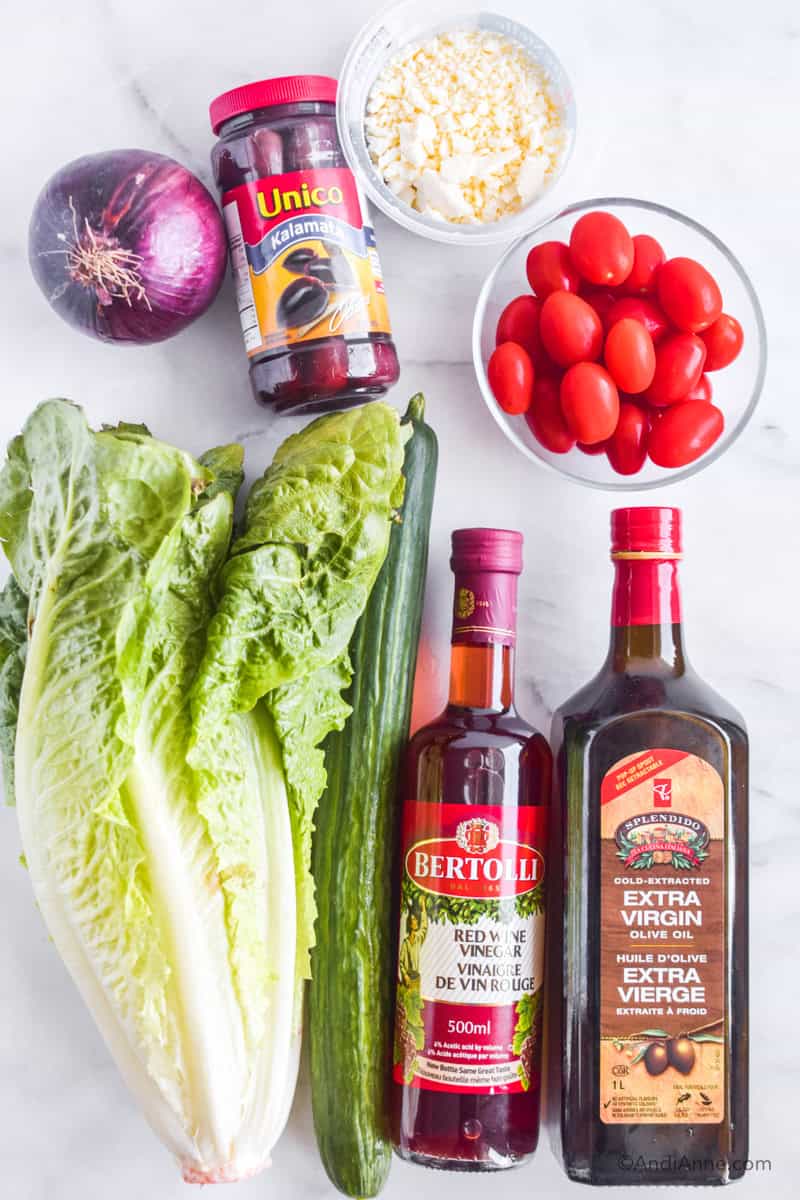 Ingredients lying down on a counter: romaine lettuce, cucumber, red wine vinegar, olive oil, bowl if grape tomatoes, container feta cheese, red onion, container of olives.