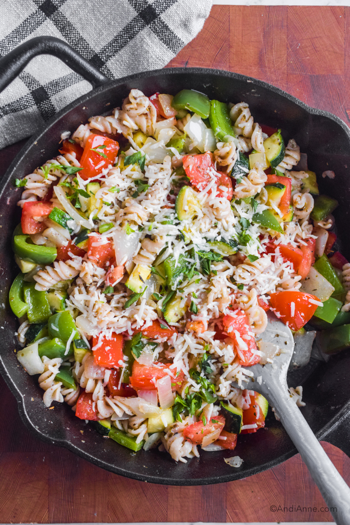 Looking down at vegetables, spiral noodles, and grated parmesan in a black skillet with a grey spatula.