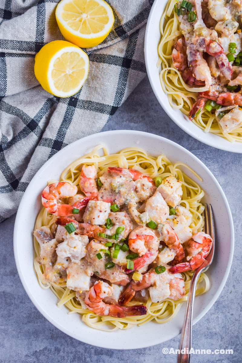 Two bowls with pasta, shrimp and cod and a fork. Kitchen towel with sliced lemon.