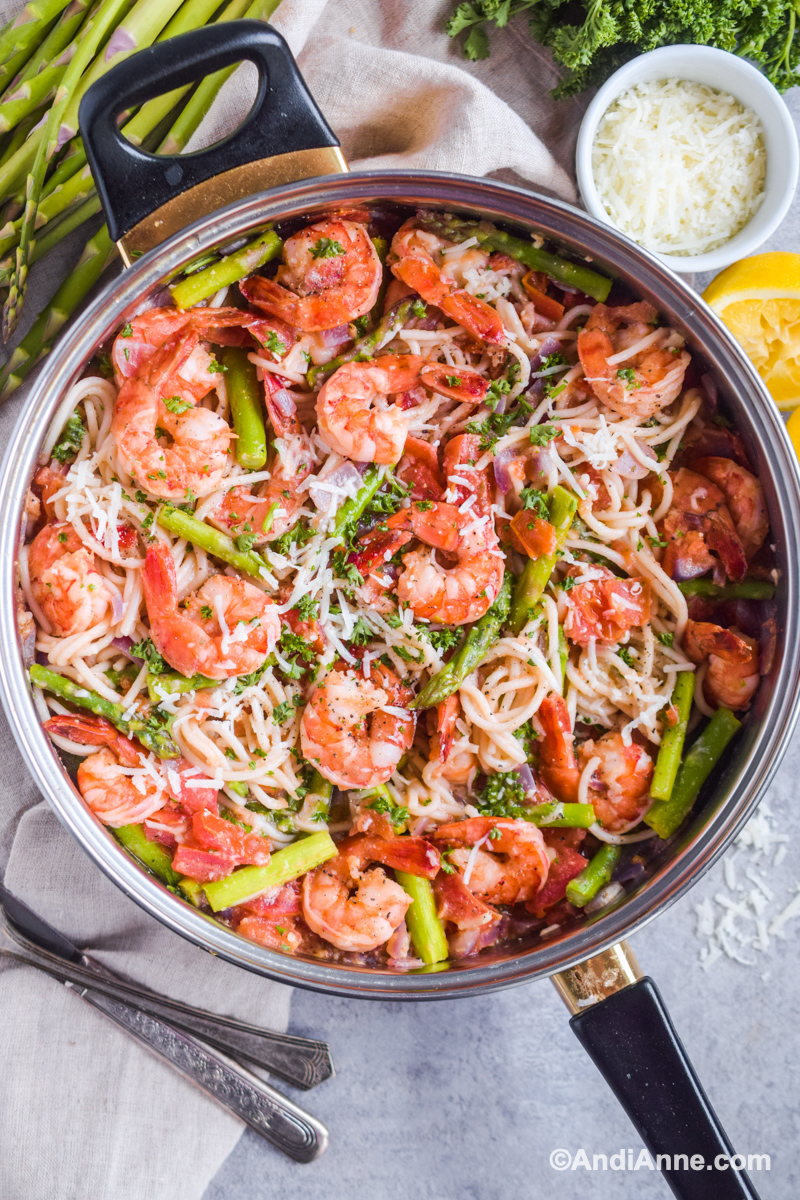 Shrimp asparagus pasta recipe in a pot surrounded with squeezed lemon slices, bowl of parmesan and asparagus spears. 