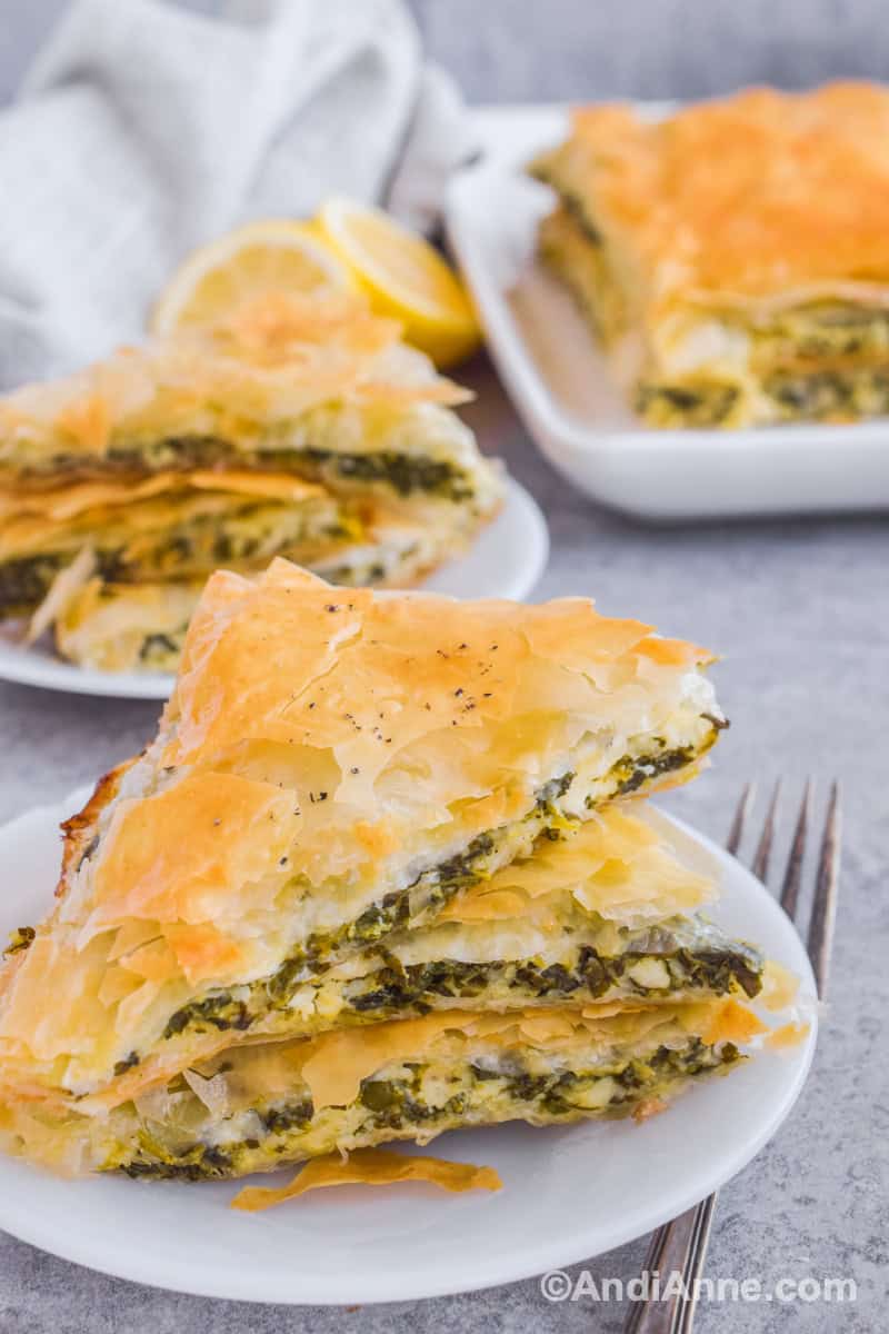 Stacked spinach feta pie on a white plate with another serving and baking dish in background.