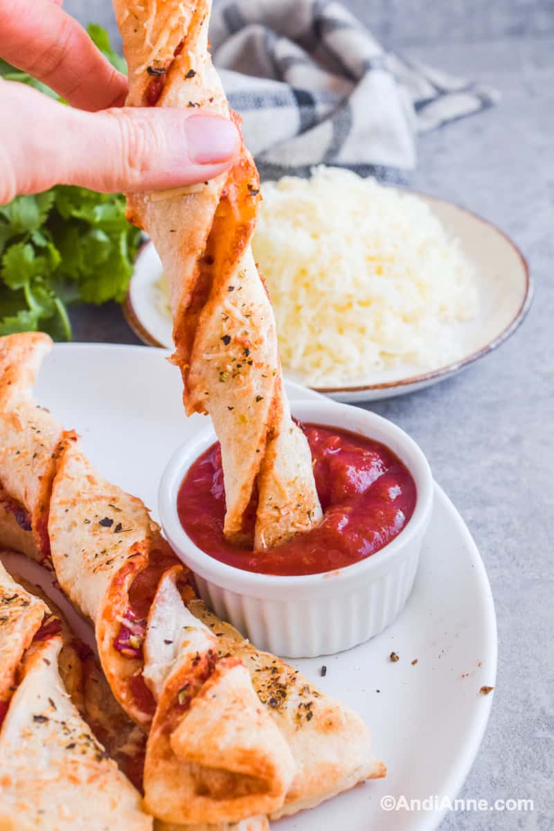 Fingers dipping the breadstick in pizza sauce with cheese and parsley in background.