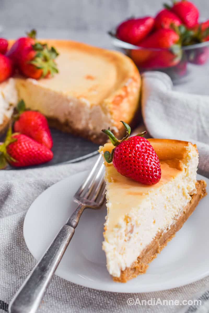 A slice of cheesecake on a plate with a fork and fresh strawberry on top. Full cheesecake and more strawberries piled in the background.