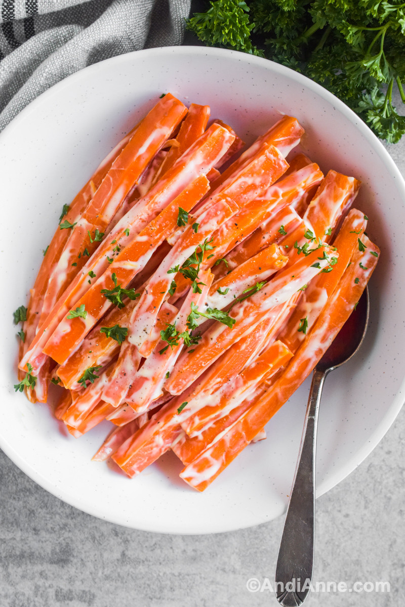 Creamy carrots in a white bowl with parsley sprinkled on top and a spoon beside.