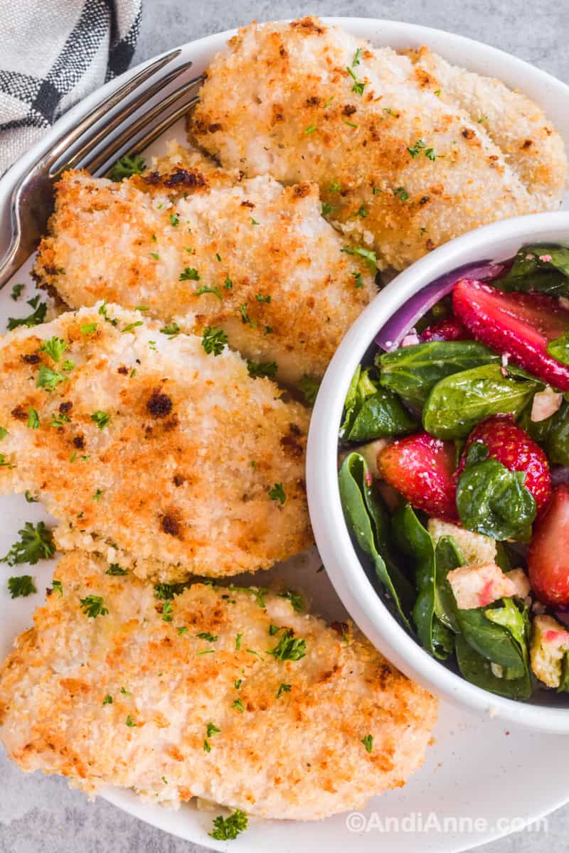 Four cooked chicken breasts on plate with bowl of salad and a fork.