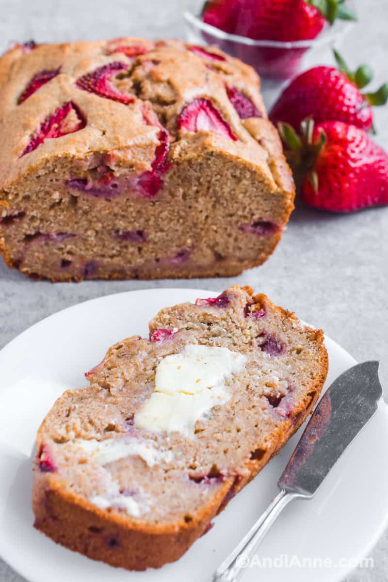 One slice of the loaf with butter and a knife on a white plate. Rest of loaf in background with fresh strawberries.