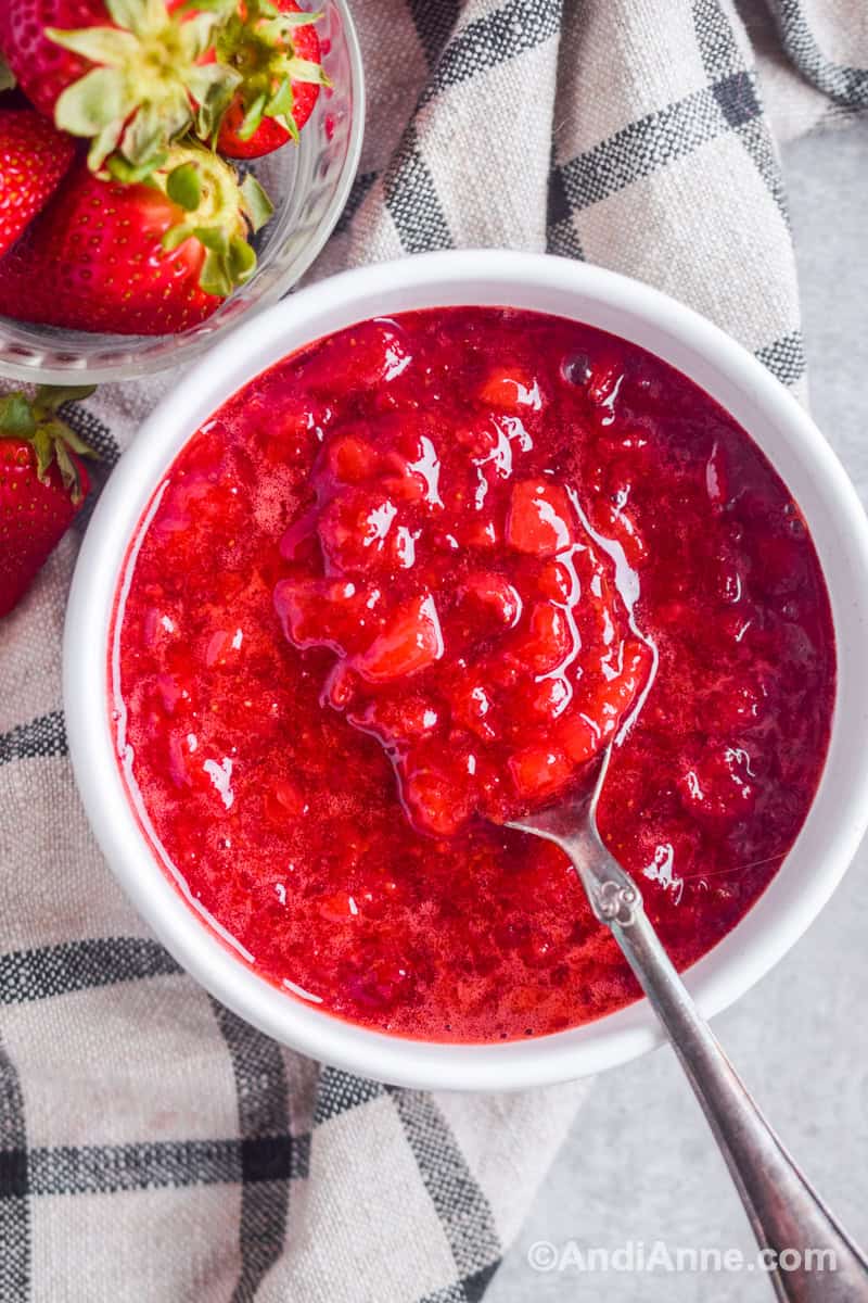 A white bowl of strawberry topping with spoon and bowl of strawberries, plus kitchen towel surrounding it.
