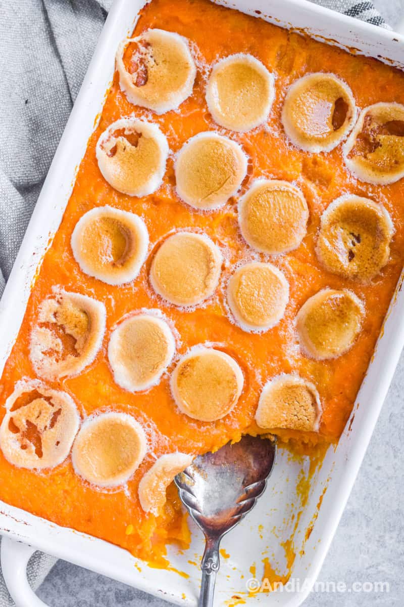 Scoop taken out of sweet potato recipe in a large white casserole dish with a large silver serving spoon. Golden brown marshmallows are on top.