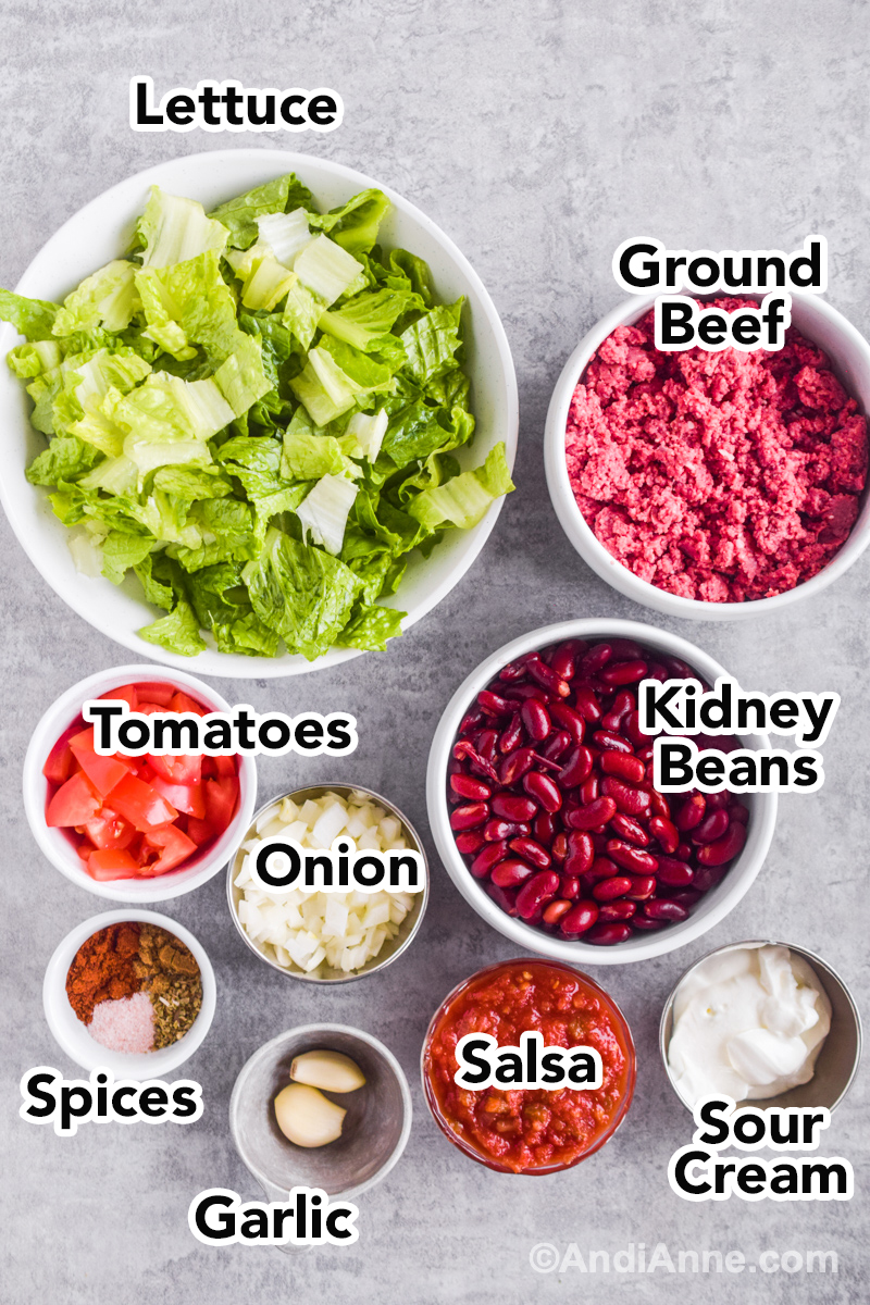 Bowls of recipe ingredients including, bowl of shredded lettuce, raw ground beef, kidney beans, chopped tomatoes, chopped onion, spices, salsa, and sour cream.