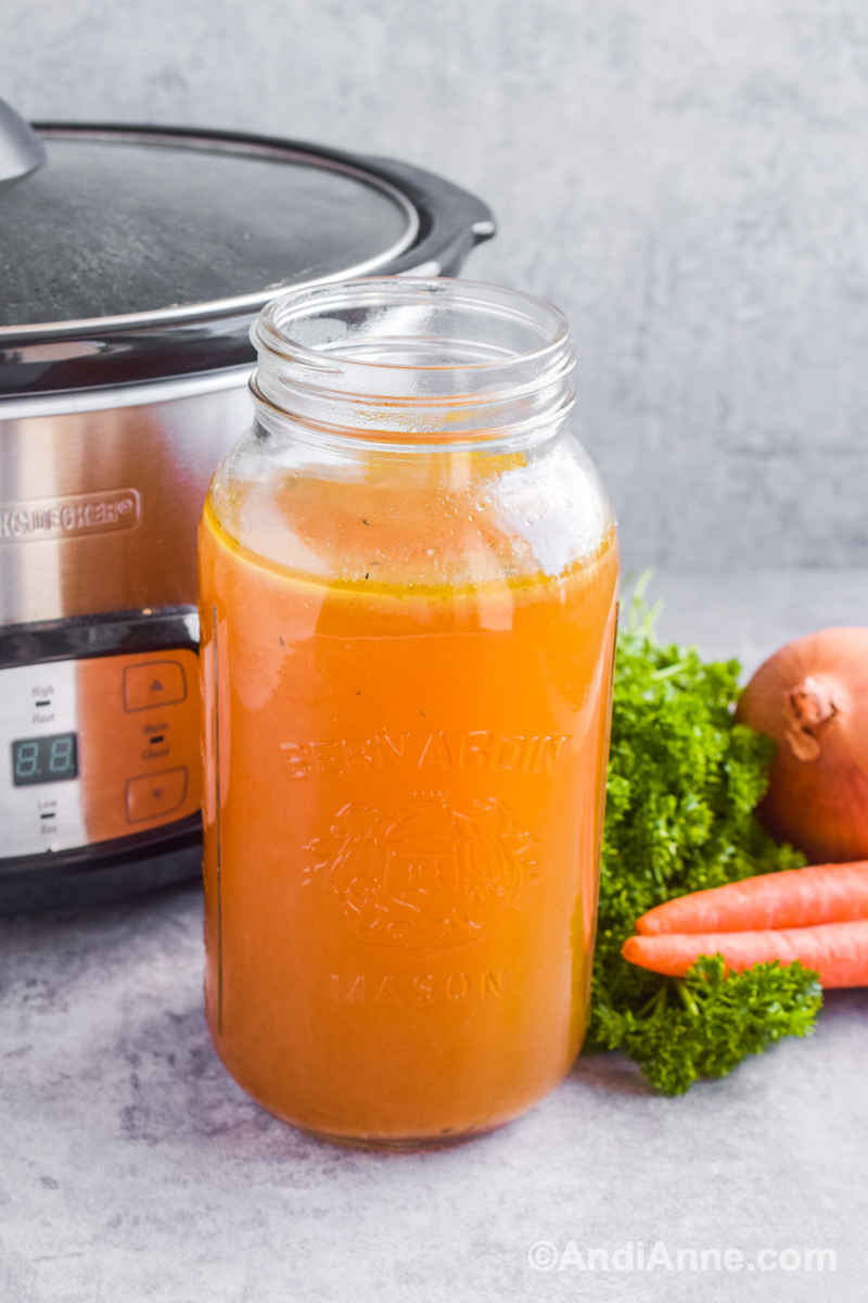 Turkey broth in large mason jar with slow cooker and fresh vegetables behind it.