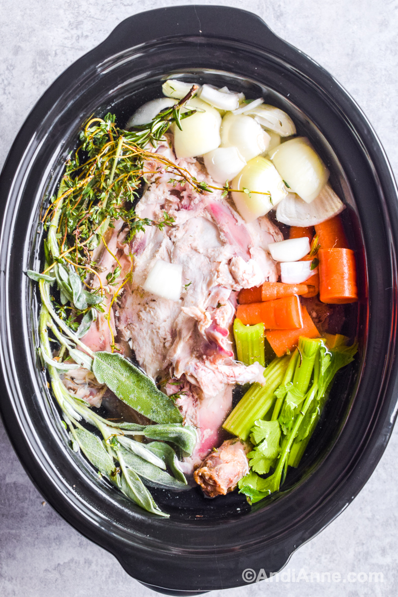 Looking down at slow cooker pot with turkey carcass, fresh herbs, onion, carrots and celery.