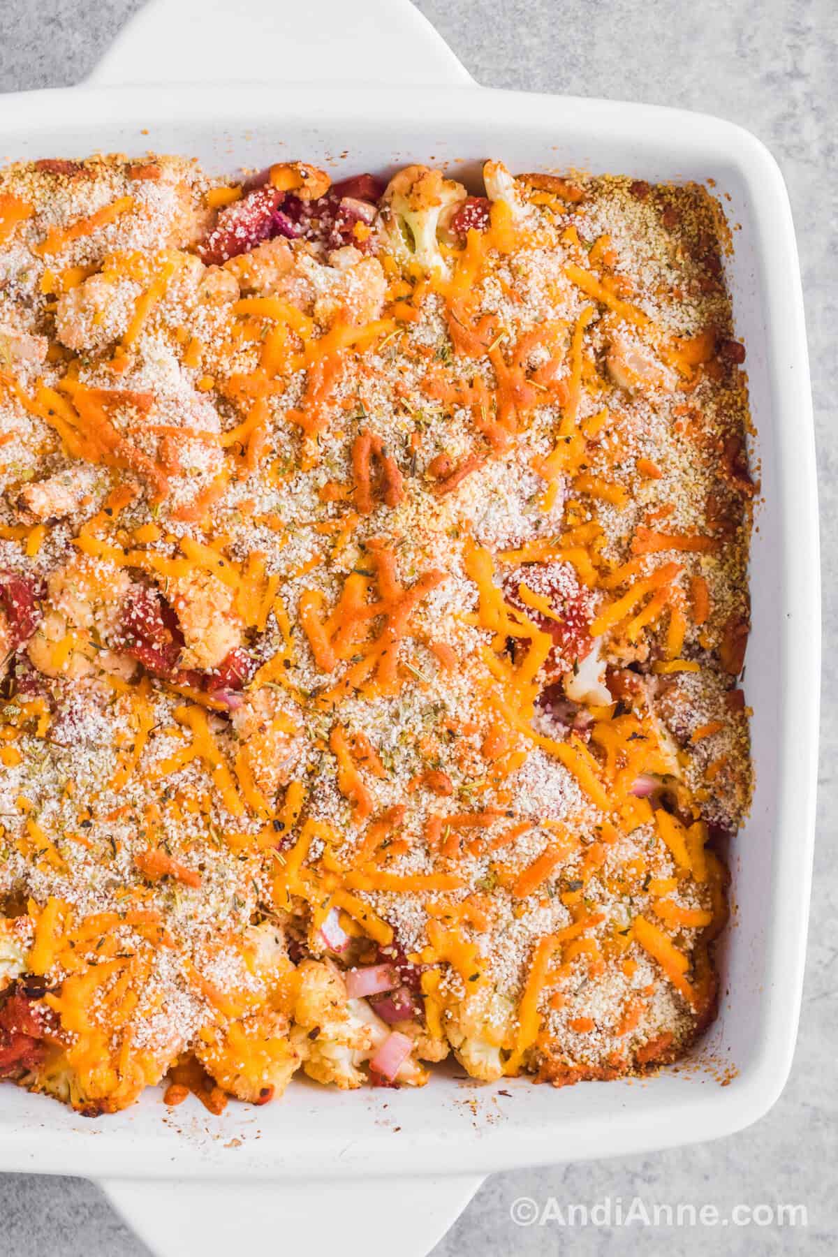 Close up of baked cheese and breadcrumbs overtop of cauliflower and tomatoes inside a white square casserole dish.