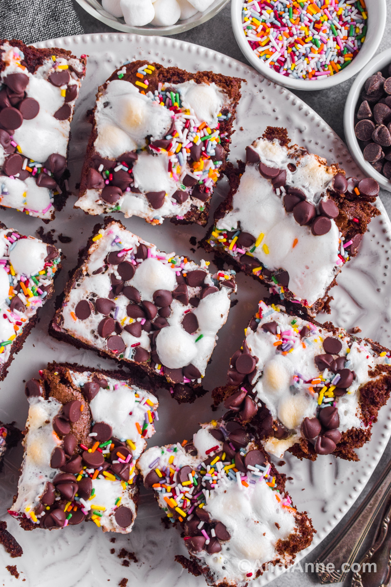 Sprinkle marshmallow cake squares on a plate with small bowls of sprinkles, chocolate chips and marshmallows beside it. 