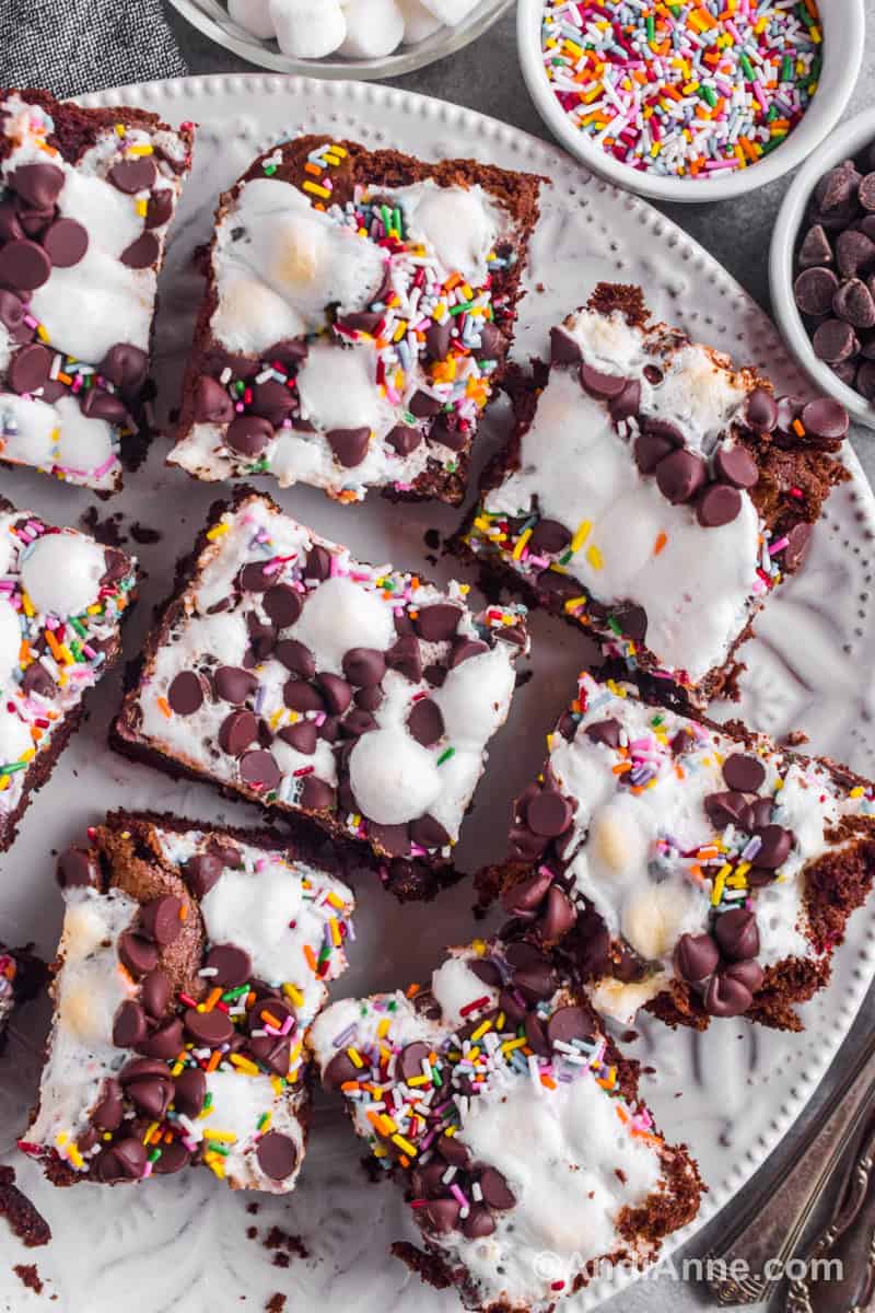Sprinkle marshmallow cake squares on a plate with small bowls of sprinkles, chocolate chips and marshmallows beside it.
