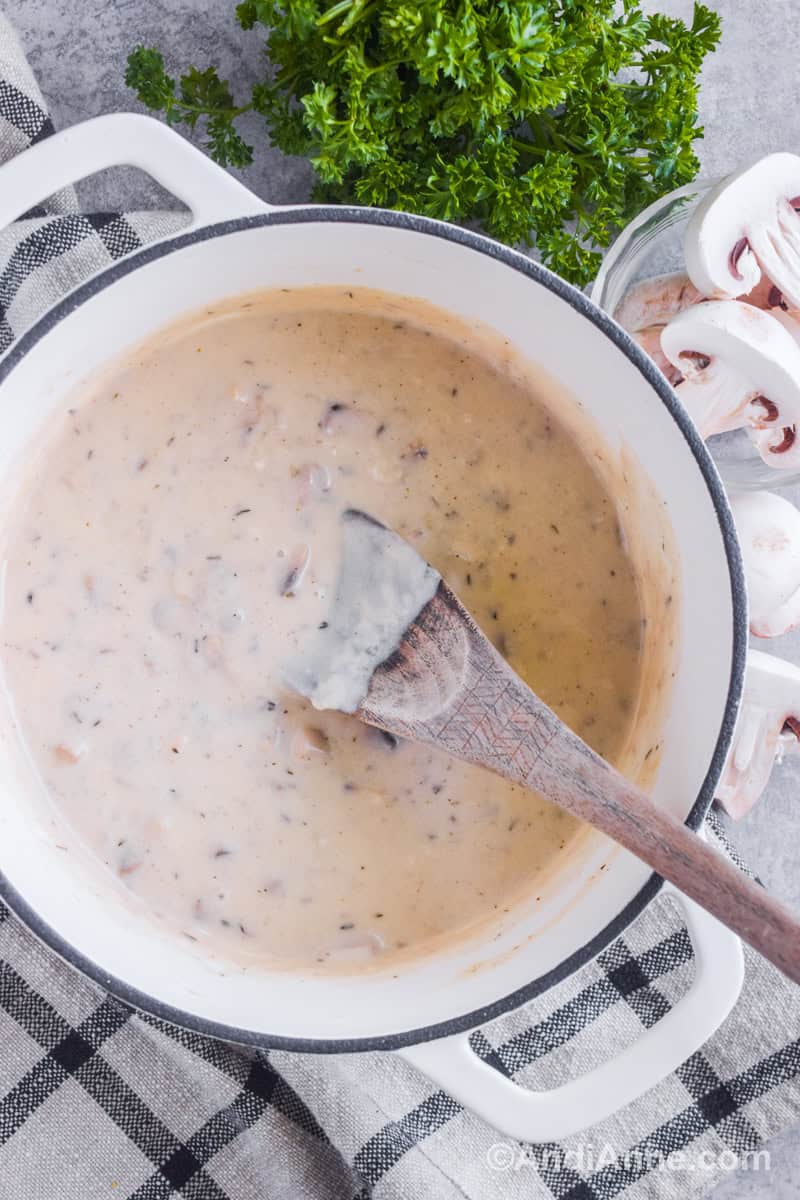 A white pot with creamy mushroom gravy and a wood spoon. Fresh parsley and chopped mushrooms surround outside of the bowl.